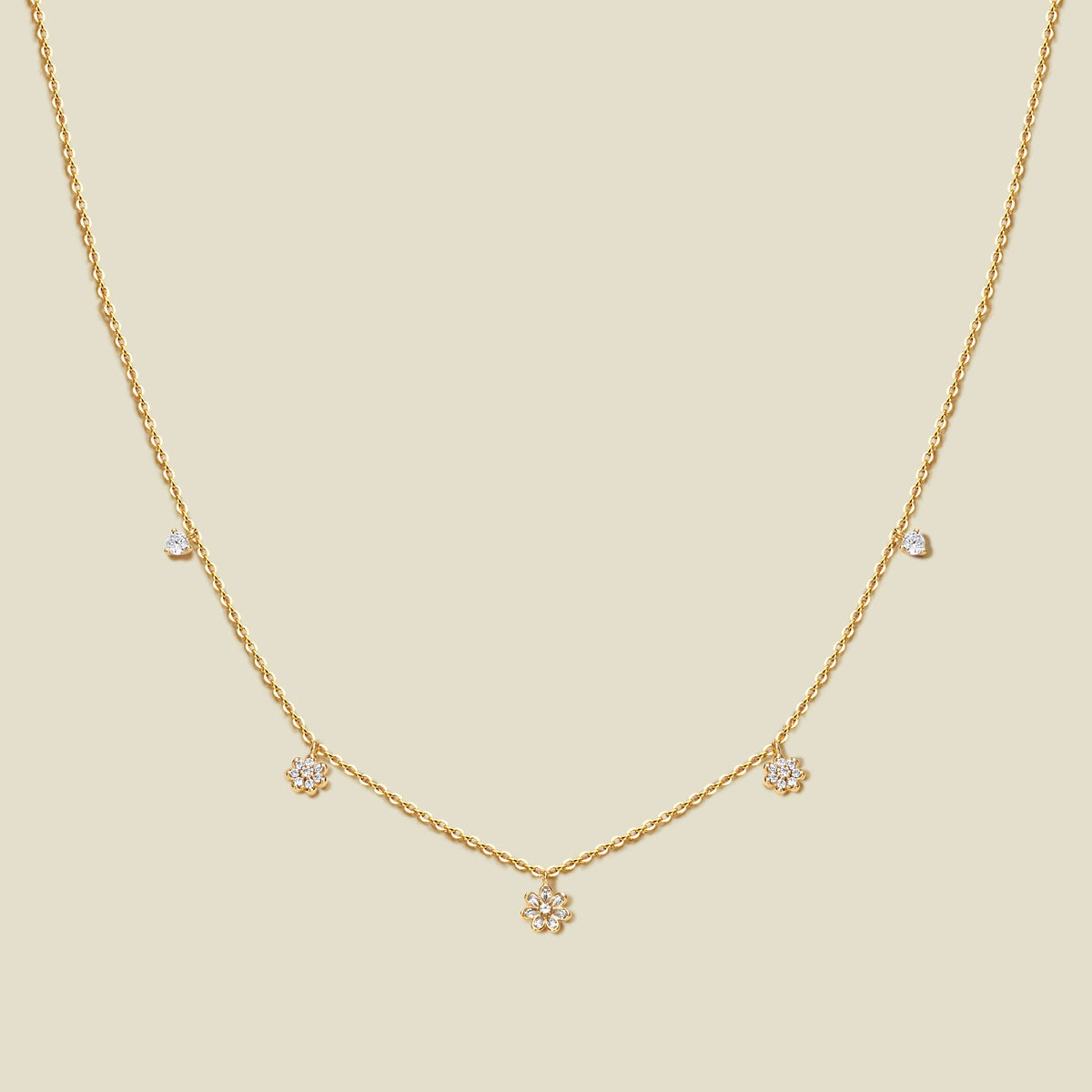 A delicate gold chain necklace with three small flowers with clear accent stones and a few tiny clear accent stones, displayed on a solid light beige background.