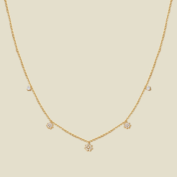 A delicate gold chain necklace with three small flowers with clear accent stones and a few tiny clear accent stones, displayed on a solid light beige background.