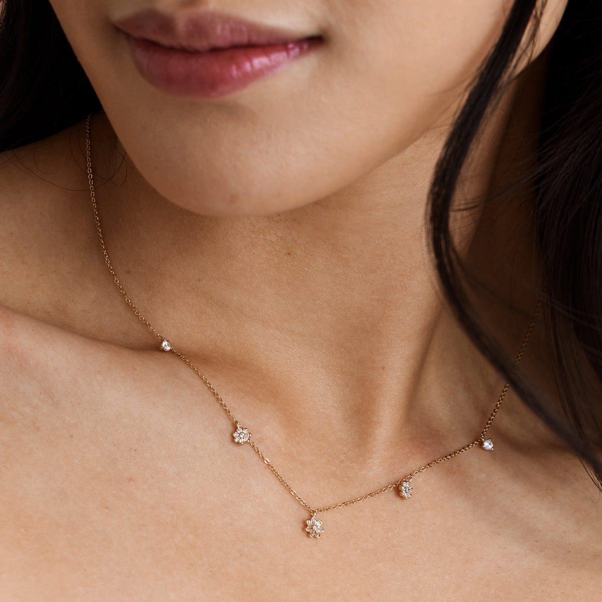 A close-up of a person's chest wearing a dainty gold chain necklace with three dangling clear gemstone flowers and two dangling round clear stones.