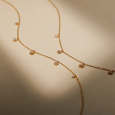 A dainty gold chain necklace featuring three dangling flowers, each with clear stones, and two round dangling clear stones on either side of them, set against a solid beige background with shadows.
