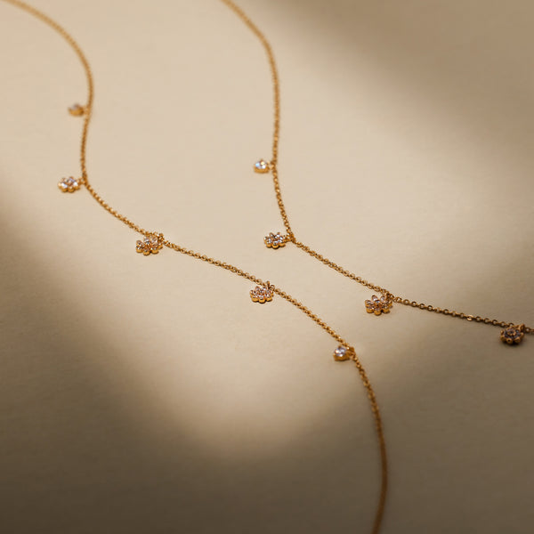 A dainty gold chain necklace featuring three dangling flowers, each with clear stones, and two round dangling clear stones on either side of them, set against a solid beige background with shadows.
