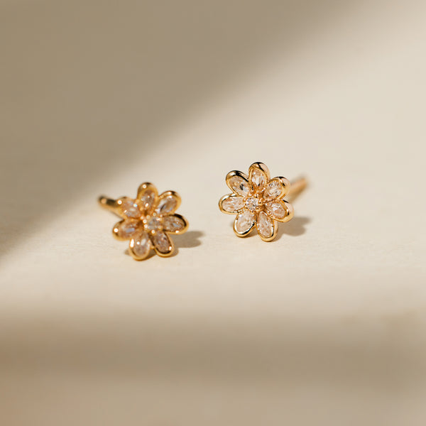 A pair of gold flower stud earrings with clear stones, displayed on a shadowed light beige background.