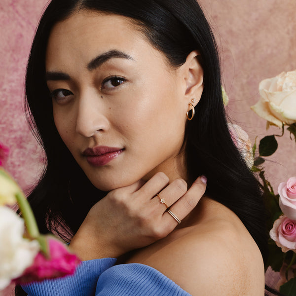 A woman in front of a light pink background surrounded by pink and white flowers, wearing multiple gold rings and earrings with clear stones.