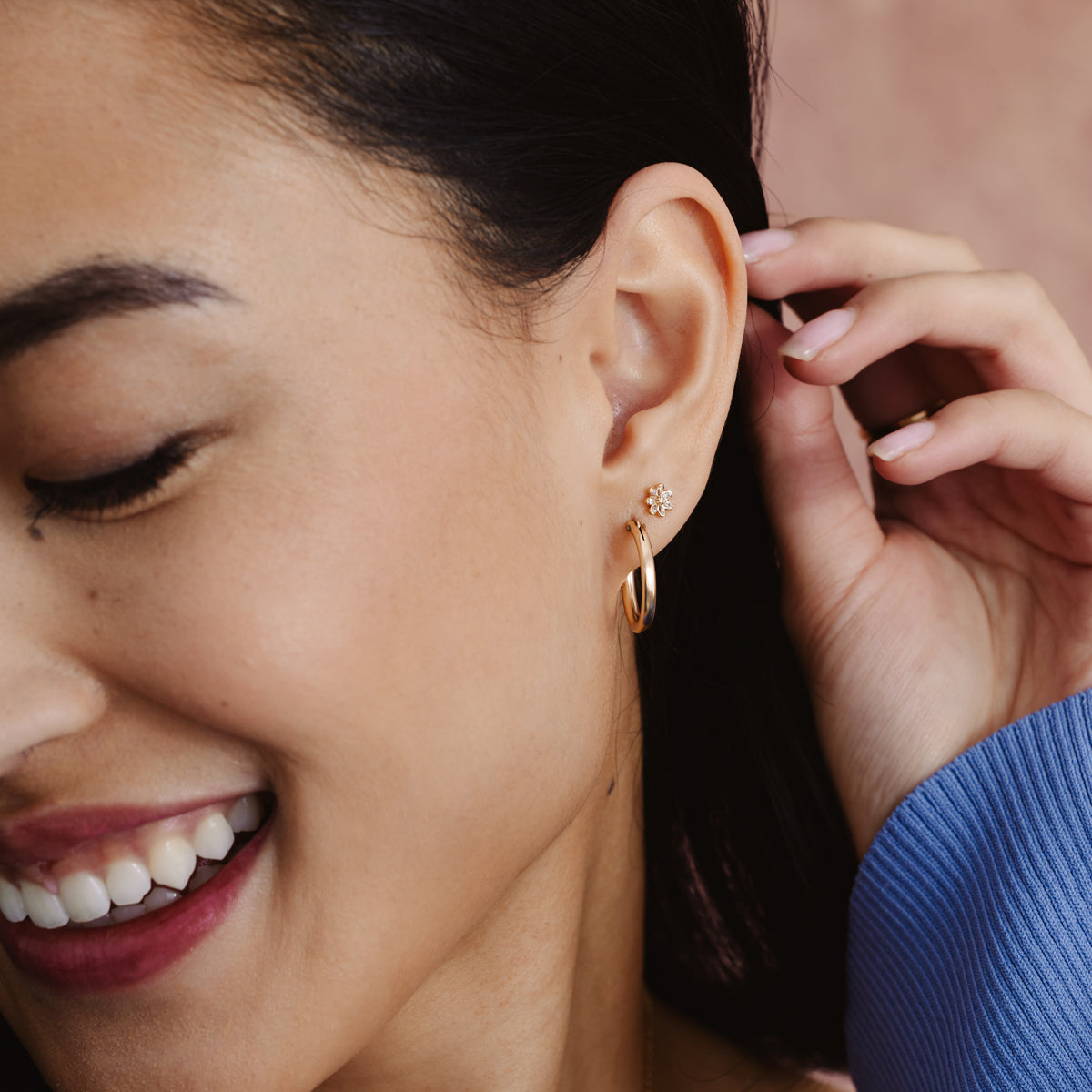 A person's ear wearing a polished gold hoop earring and a gold flower stud earring with clear stones.