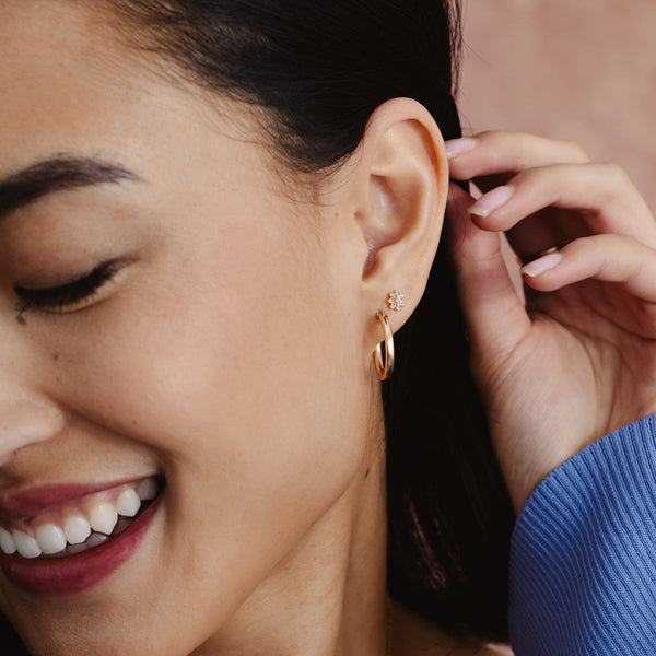 A person's ear wearing a polished gold hoop earring and a gold flower stud earring with clear stones.