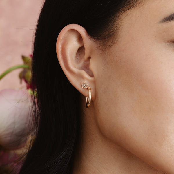 A person's ear wearing a polished gold hoop earring and a gold flower stud earring with clear stones.