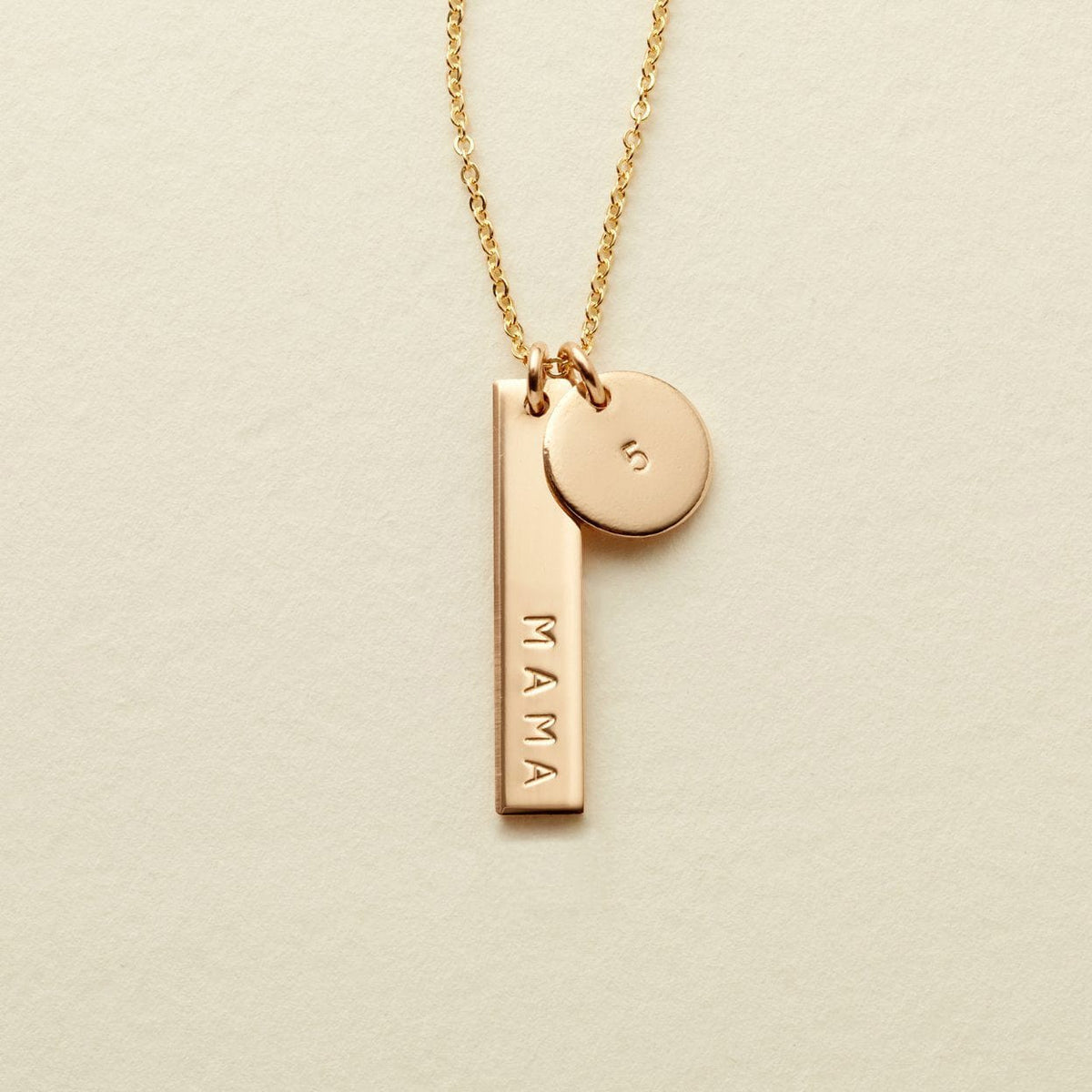 A delicate gold necklace featuring a bar pendant engraved with the name 'MAMA' and a small round disc charm stamped with the number '5', displayed on a textured off-white background.