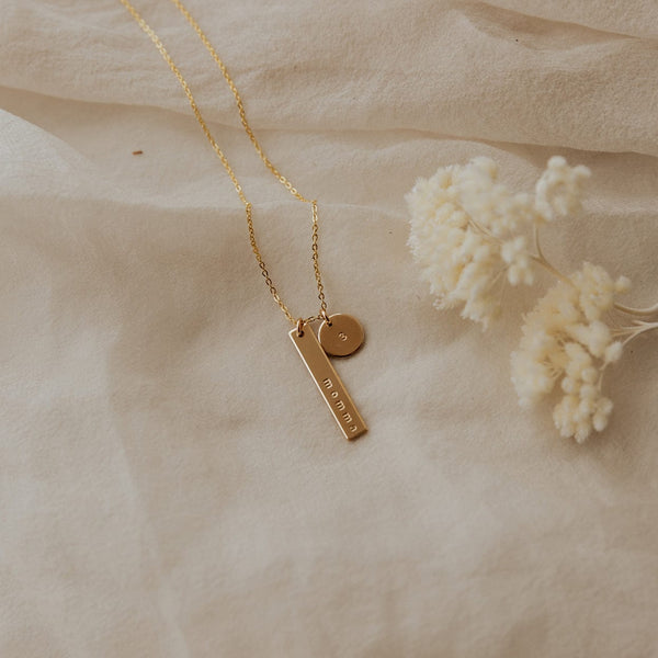 A delicate gold pendant chain necklace with a vertical bar charm engraved with the word 'momma' and a round disc charm engraved with the number '3'. The necklace is laying on an off-white linen fabric with a small sprig of round white and beige foliage beside it.