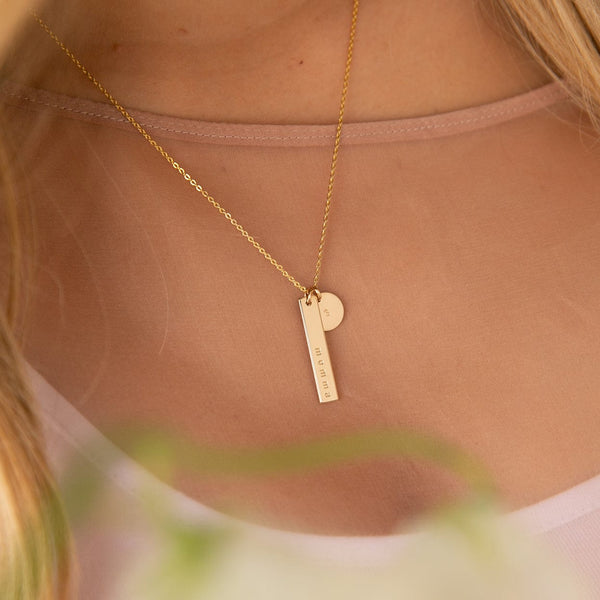 A close-up of a person wearing a high neck blush pink mesh top with a delicate gold pendant chain necklace. The necklace has a vertical bar charm engraved with the word 'momma' and a small round disc charm engraved with the number '3'.