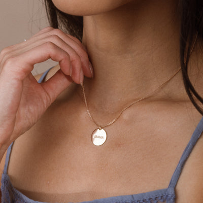 A close-up of a woman wearing a delicate gold chain necklace with a round gold pendant engraved with the word 'MAMA'.