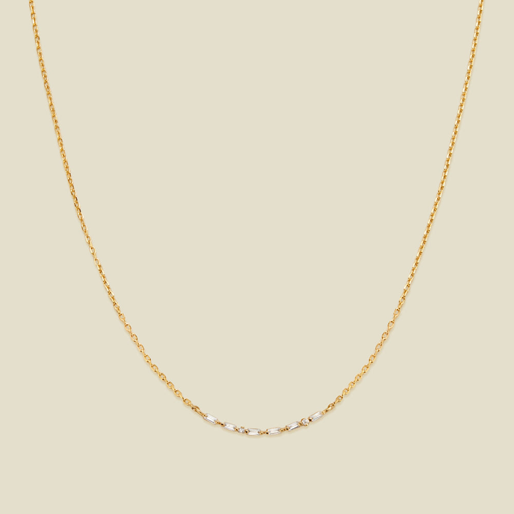 A delicate gold chain necklace featuring a short central segment of clear round and baguette-cut gemstones, displayed against a solid light beige background.