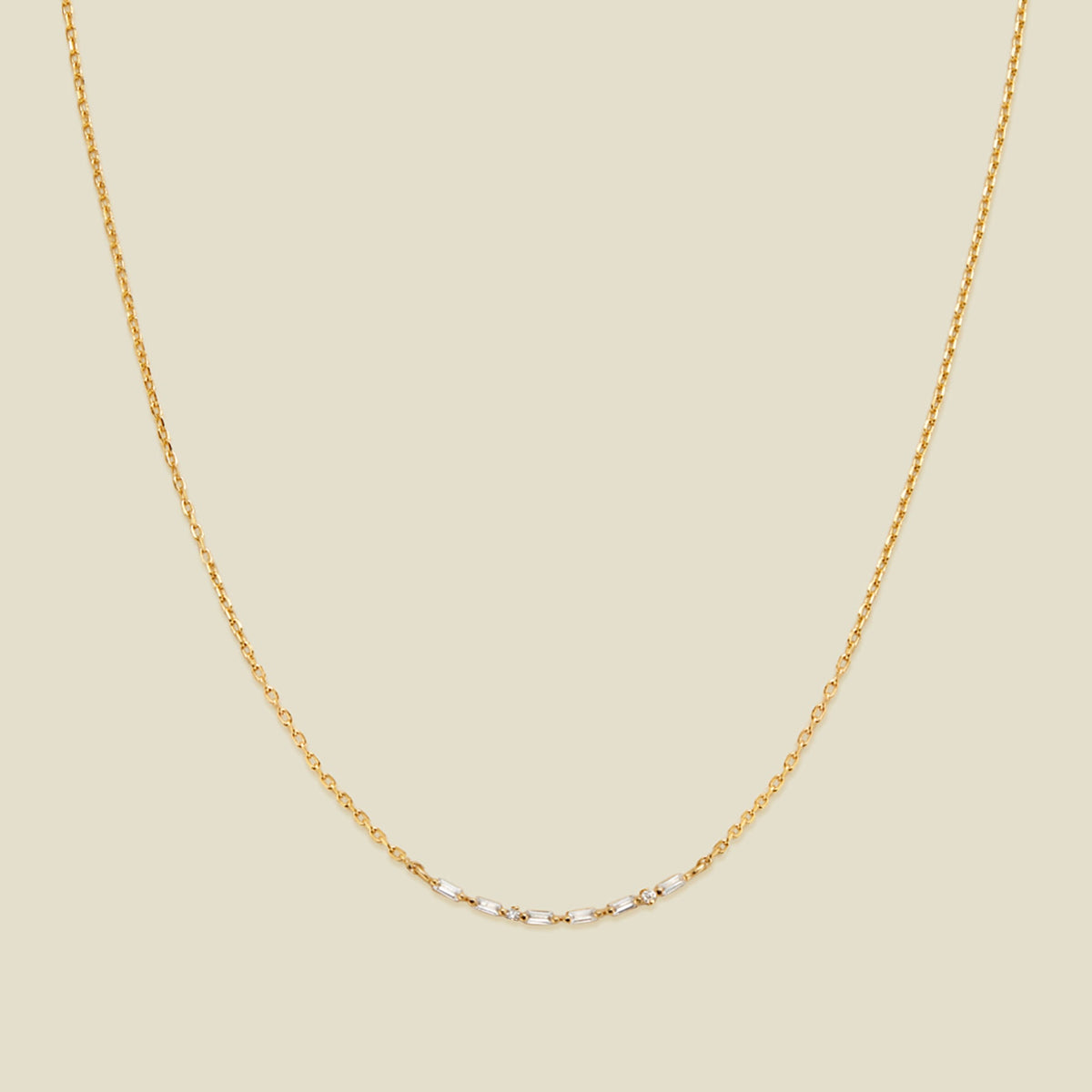 A delicate gold chain necklace featuring a short central segment of clear round and baguette-cut gemstones, displayed against a solid light beige background.
