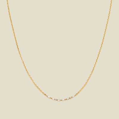 A delicate gold chain necklace featuring a short central segment of clear round and baguette-cut gemstones, displayed against a solid light beige background.