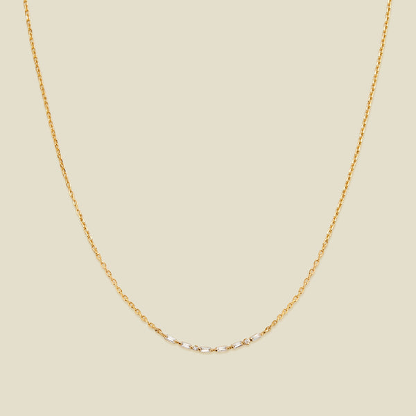 A delicate gold chain necklace featuring a short central segment of clear round and baguette-cut gemstones, displayed against a solid light beige background.