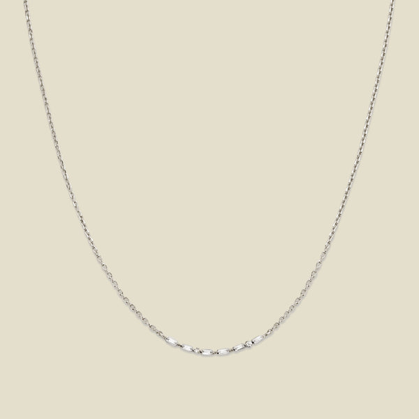 A delicate silver chain necklace featuring a short central segment of clear round and baguette-cut gemstones, displayed against a solid light beige background.