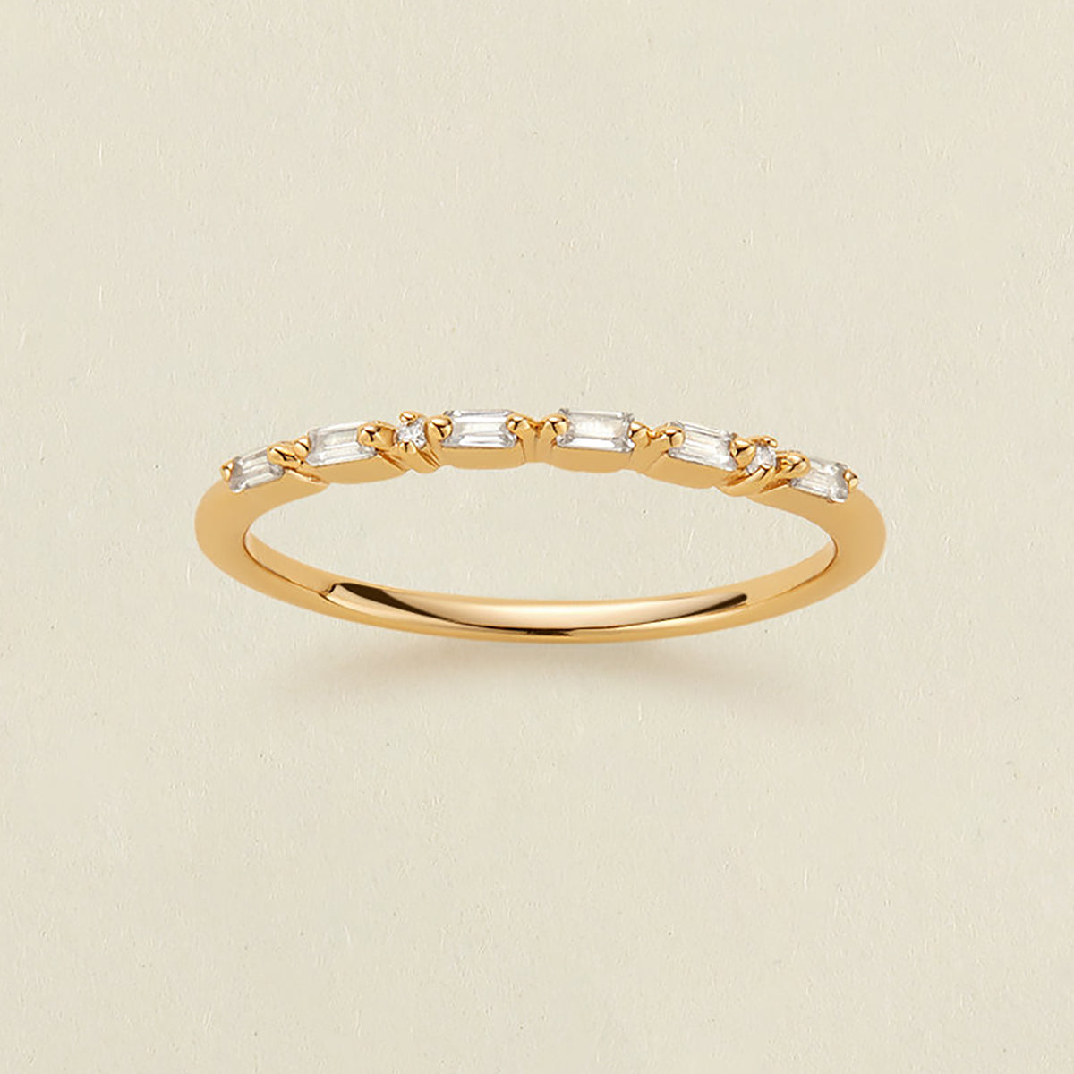 A delicate yellow gold ring featuring a row of baguette-cut and round clear stones in a morse code pattern, displayed on a light beige textured background.