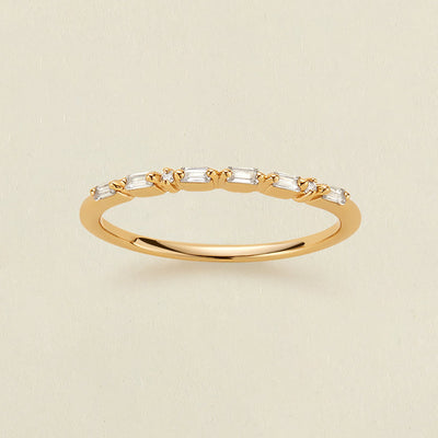 A delicate yellow gold ring featuring a row of baguette-cut and round clear stones in a morse code pattern, displayed on a light beige textured background.