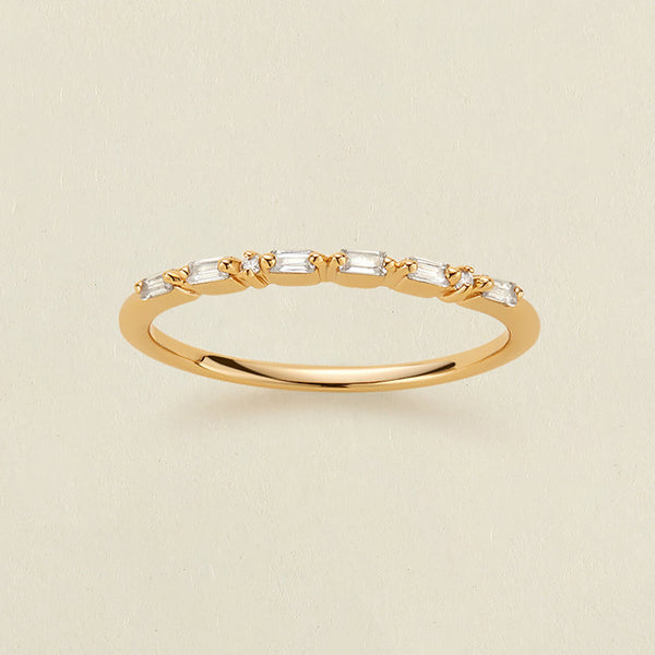 A delicate yellow gold ring featuring a row of baguette-cut and round clear stones in a morse code pattern, displayed on a light beige textured background.