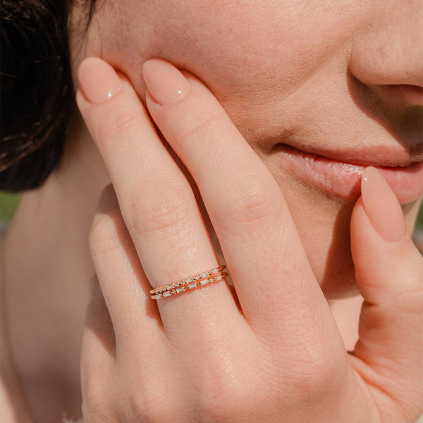 A close-up of a hand resting on someone's face showcasing two gold rings. Each ring has a different pattern of baguette and round cut clear gemstones in a morse code pattern.
