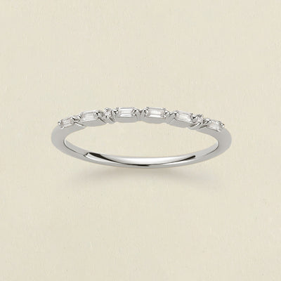 A delicate silver ring featuring a row of baguette-cut and round clear stones in a morse code pattern, displayed on a light beige textured background.