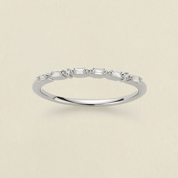 A delicate silver ring featuring a row of baguette-cut and round clear stones in a morse code pattern, displayed on a light beige textured background.