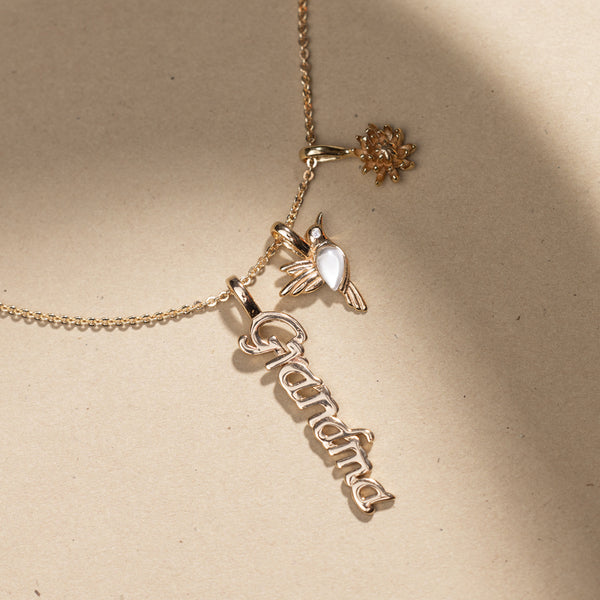 Gold pendant chain necklace with 'Grandma', hummingbird and chrysanthemum charm pendants on a beige background
