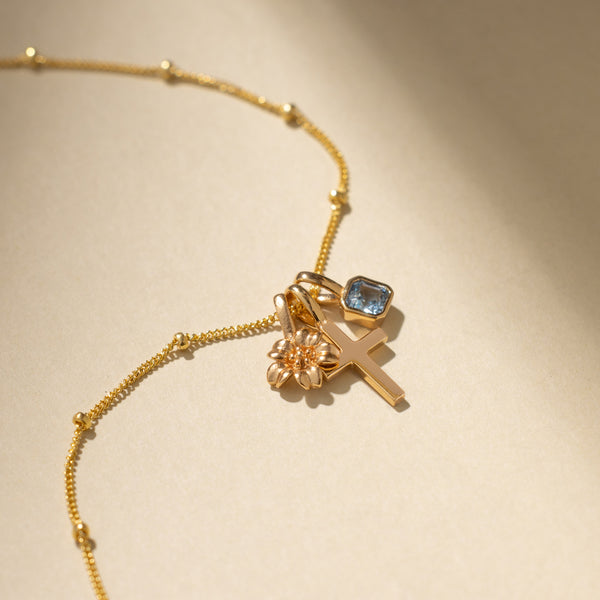 A gold chain necklace with small round stationary beads, holding a small gold cherry blossom flower charm, a cross charm, and a square light-blue gemstone charm.