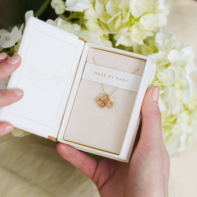 A person holding open a small cream colored jewelry box with a gold pendant chain inside. The chain has three small flower charms on it.