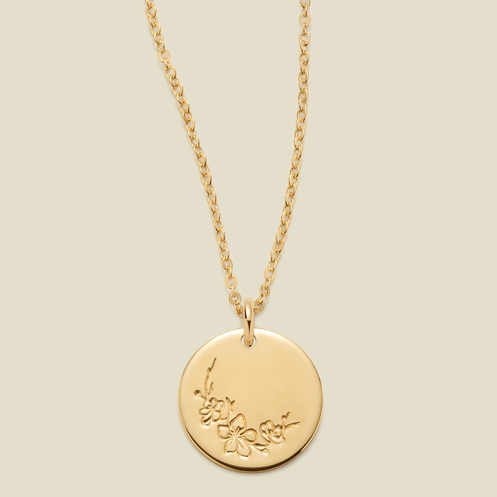 A gold chain necklace with a round gold disc pendant featuring an engraved cherry blossom flower design, displayed against a solid light beige background.