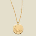 A gold chain necklace with a round gold disc pendant featuring an engraved cherry blossom flower design, displayed against a solid light beige background.