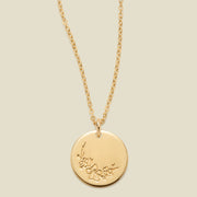 A gold chain necklace with a round gold disc pendant featuring an engraved cherry blossom flower design, displayed against a solid light beige background.