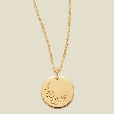 A gold chain necklace with a round gold disc pendant featuring an engraved cherry blossom flower design, displayed against a solid light beige background.