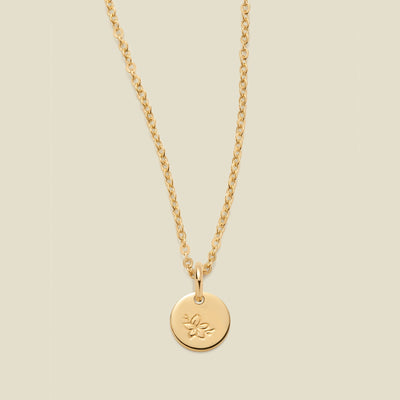 A gold chain necklace with a round gold disc pendant featuring an engraved cherry blossom flower design, displayed against a solid light beige background.