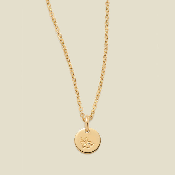A gold chain necklace with a round gold disc pendant featuring an engraved cherry blossom flower design, displayed against a solid light beige background.