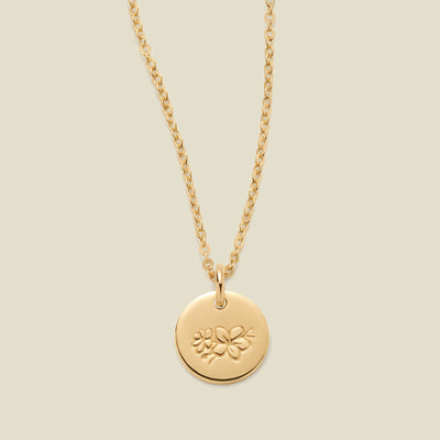 A gold chain necklace with a round gold disc pendant featuring an engraved cherry blossom flower design, displayed against a solid light beige background.