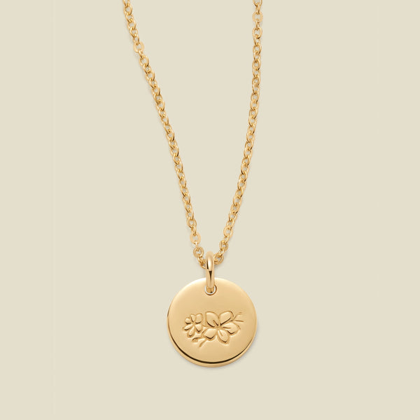 A gold chain necklace with a round gold disc pendant featuring an engraved cherry blossom flower design, displayed against a solid light beige background.