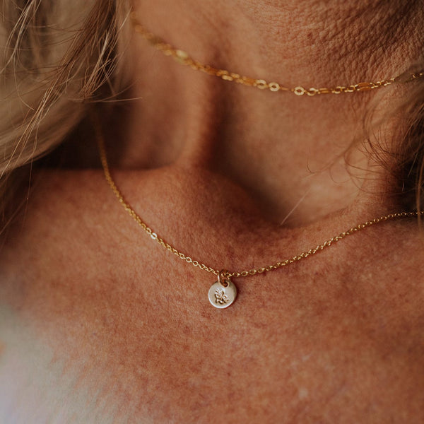 A close-up of a person wearing a delicate gold pendant chain with a flat round engraved disc charm. The engraving is of a cherry blossom flower.