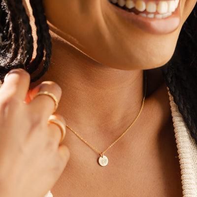 A close-up of a woman wearing a delicate gold pendant chain with a flat round disc. The disc is engraved with a cherry blossom floral design.