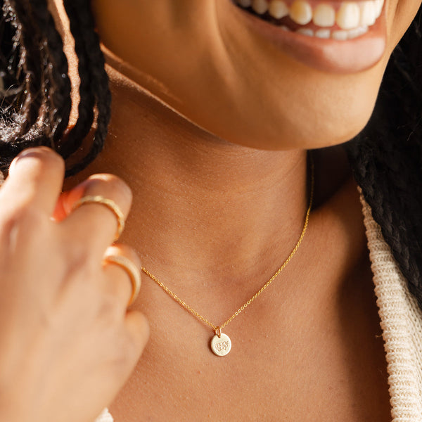 A close-up of a woman wearing a delicate gold pendant chain with a flat round disc. The disc is engraved with a cherry blossom floral design.