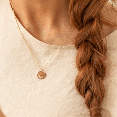 A close-up of a woman with red hair in a braid, wearing a white top and a delicate gold pendant chain necklace featuring a flat round disc charm. The charm is engraved with a cherry blossom floral design.