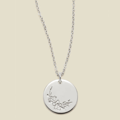 A silver chain necklace with a round silver disc pendant featuring an engraved cherry blossom flower design, displayed against a solid light beige background.