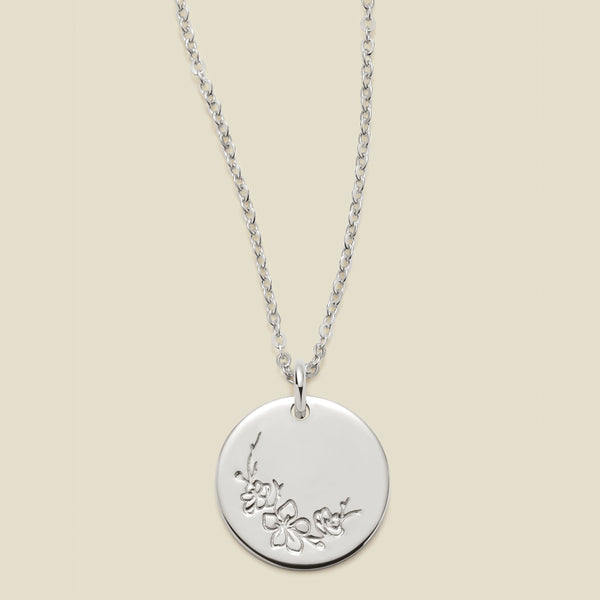 A silver chain necklace with a round silver disc pendant featuring an engraved cherry blossom flower design, displayed against a solid light beige background.