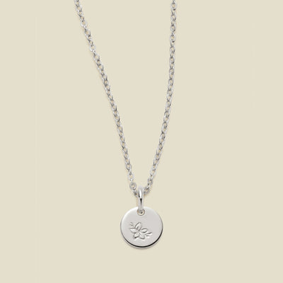 A silver chain necklace with a round silver disc pendant featuring an engraved cherry blossom flower design, displayed against a solid light beige background.