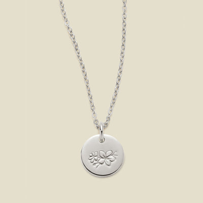 A silver chain necklace with a round silver disc pendant featuring an engraved cherry blossom flower design, displayed against a solid light beige background.