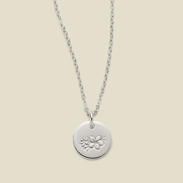 A silver chain necklace with a round silver disc pendant featuring an engraved cherry blossom flower design, displayed against a solid light beige background.