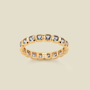 A gold eternity ring featuring a continuous row of square cut light blue gemstones, shown on a solid light beige background.