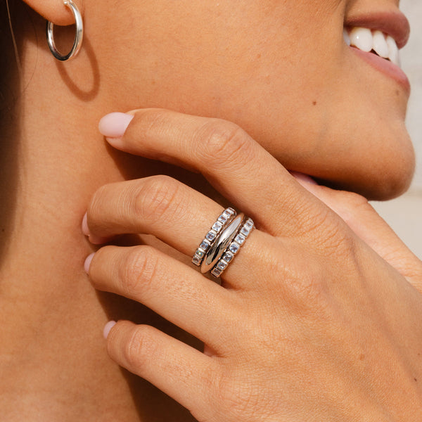 A person's hand resting on their neck wearing three stacked silver rings. One is a polished bubble ring and the other two are eternity style rings, each with light blue square cut gemstones in a single continuous row.