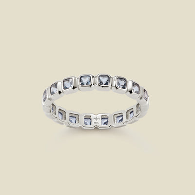 A silver eternity ring featuring a continuous row of square cut light blue gemstones, shown on a solid light beige background.