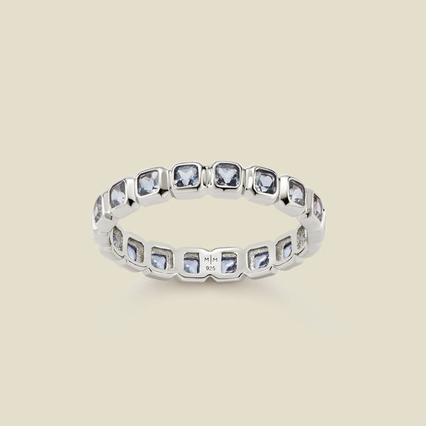 A silver eternity ring featuring a continuous row of square cut light blue gemstones, shown on a solid light beige background.