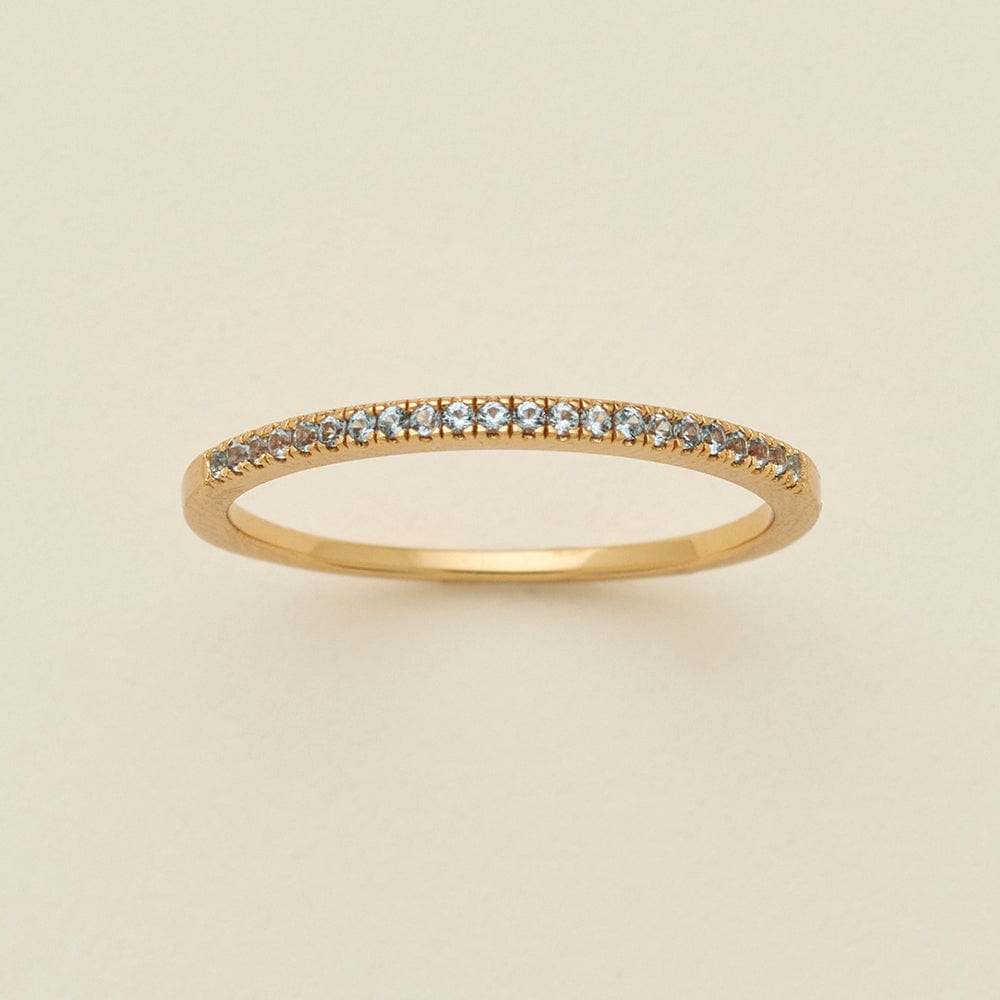 A gold ring featuring a single row of small round-cut light blue-grey gemstones, photographed on a clean, solid, warm off-white background.